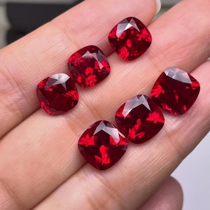 Könnte beinhalten: Sechs facettierte Edelsteine im Kissenschliff in tiefroter Farbe. Die Edelsteine werden in einer Hand gehalten, was ihre Klarheit und Brillanz zeigt. Die quadratische Form und die satte Farbe deuten auf Rubine oder Granate hin.