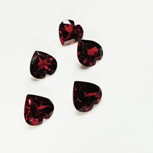 Puede incluir: Cinco gemas facetadas en forma de corazón, de un tono rojo intenso, están dispersas sobre una superficie blanca. Las gemas tienen un corte brillante, captando la luz y mostrando reflejos internos. Ideales para la fabricación de joyas.