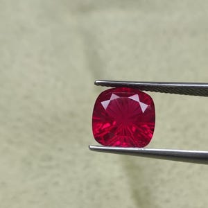 May include: A faceted, cushion-cut ruby gemstone held by tweezers. The ruby is a vibrant red colour with internal light reflections. The gemstone is square with rounded corners.