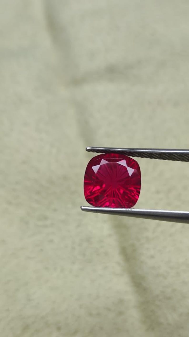 May include: A faceted, cushion-cut ruby gemstone held by tweezers. The ruby is a vibrant red colour with internal light reflections. The gemstone is square with rounded corners.