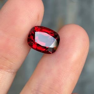 Puede incluir: Una gema roja oscura facetada de forma rectangular. La gema tiene una superficie pulida con múltiples facetas que reflejan la luz, creando un brillo brillante. El color es un rojo rico y saturado, con toques de tonos más oscuros.