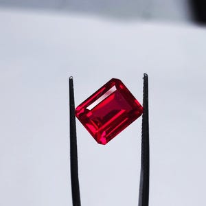 May include: A deep red gemstone with a rectangular emerald cut, held by tweezers against a white background.
