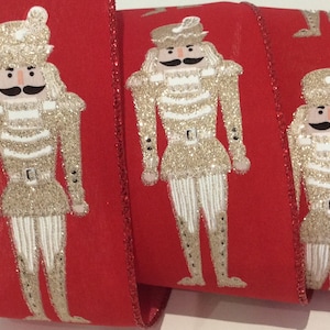 May include: Red ribbon with a repeating pattern of gold glitter nutcracker figures. The nutcrackers have black moustaches, white striped trousers, and wear hats. The ribbon's edges are trimmed with a glittery red border, suitable for festive crafts and decorations.