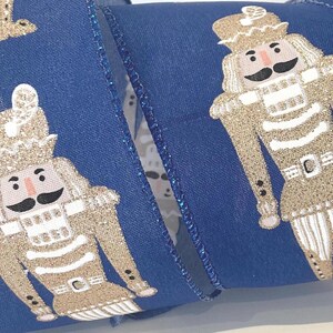May include: Royal blue ribbon with a repeating pattern of gold glitter nutcracker soldier figures. The nutcrackers have black moustaches and wear gold hats and uniforms. The ribbon is suitable for festive crafts and decorations.