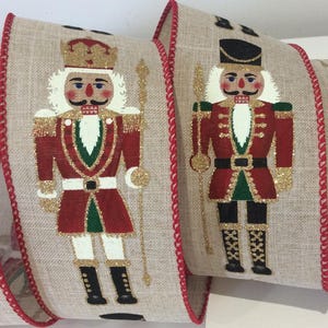 May include: Two rolls of decorative ribbon with a repeating pattern of nutcracker soldiers. The nutcrackers are in red, green, and gold uniforms with white hair and moustaches. The ribbon has a beige background with red trim.