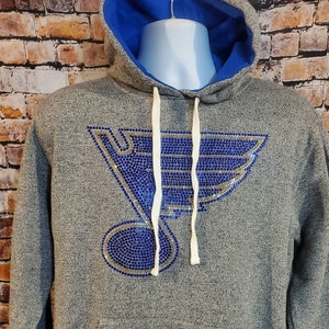 May include: Gray hoodie with a blue interior and a large blue and gold emblem on the front. The hoodie has a drawstring hood, a front pocket, and blue and white striped trim on the cuffs and hem.