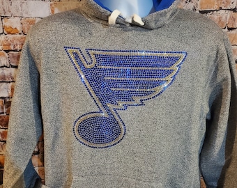 May include: Gray hoodie with a blue and gold rhinestone logo. The hoodie has a blue hood lining, white drawstrings, and a blue and white striped hem. The pockets and cuffs have white and blue accents. A tag is attached to the pocket.