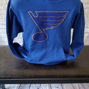 May include: Royal blue hoodie with a large, rhinestone-encrusted hockey team logo on the front. The logo is a blue and gold note with a wing design.
