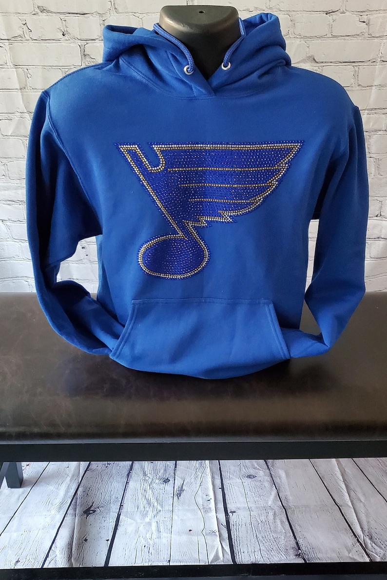May include: Royal blue hoodie with a large, rhinestone-encrusted hockey team logo on the front. The logo is a blue and gold note with a wing design.