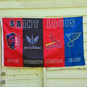 Puede incluir: Una bandera roja, negra y azul con las palabras "SAINT LOUIS" impresas en la parte superior. La bandera está dividida en cuatro secciones, cada una con el logotipo de un equipo deportivo diferente: City SC, St. Louis Battlehawks, St. Louis Cardinals y St. Louis Blues.