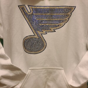 May include: White hoodie with a blue and gold rhinestone design of a hockey team logo. The logo is a stylized bird with a musical note.