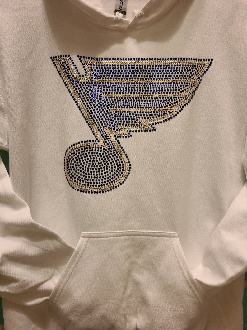 May include: White hoodie with a blue and gold rhinestone design of a hockey team logo. The logo is a stylized bird with a musical note.
