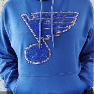 May include: A royal blue hoodie with a large rhinestone design of a hockey team logo. The logo is a stylized bird with a musical note in the center. The rhinestones are blue, gold, and clear.