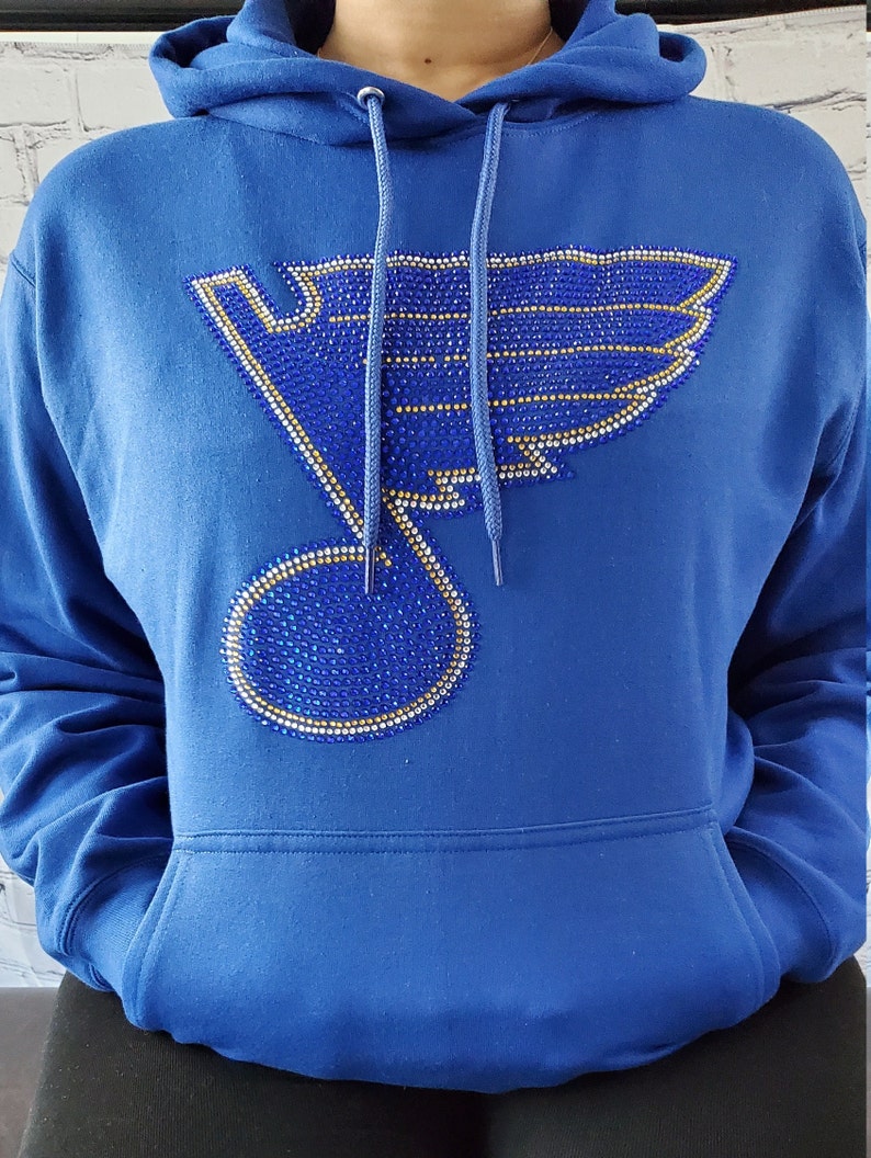 May include: A royal blue hoodie with a large rhinestone design of a hockey team logo. The logo is a stylized bird with a musical note in the center. The rhinestones are blue, gold, and clear.