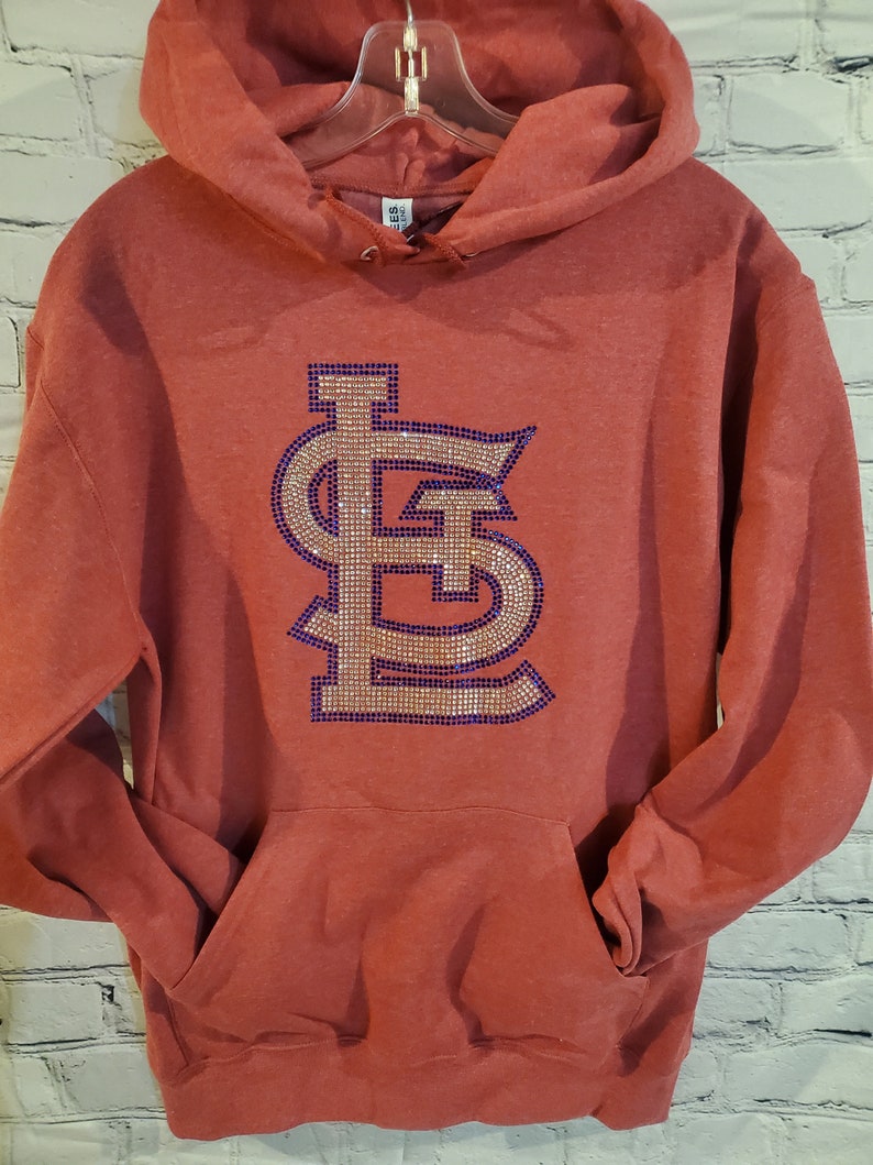 May include: Red hooded sweatshirt with a large rhinestone-embellished "LS" logo in gold and purple.