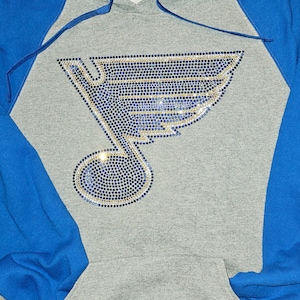 May include: Gray hoodie with blue sleeves and hood lining. The front features a rhinestone design of a musical note and a stylized wing, in blue and gold. The hoodie has a front pocket and a drawstring.