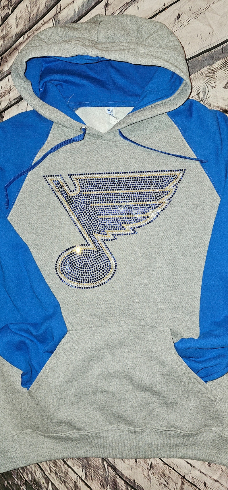 May include: Gray hoodie with blue sleeves and hood lining. The front features a rhinestone design of a musical note and a stylized wing, in blue and gold. The hoodie has a front pocket and a drawstring.