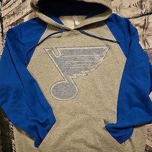 May include: Gray hoodie with blue sleeves and a hood. The front features a rhinestone design of the St. Louis Blues logo. The hoodie has a front pocket and a drawstring hood.
