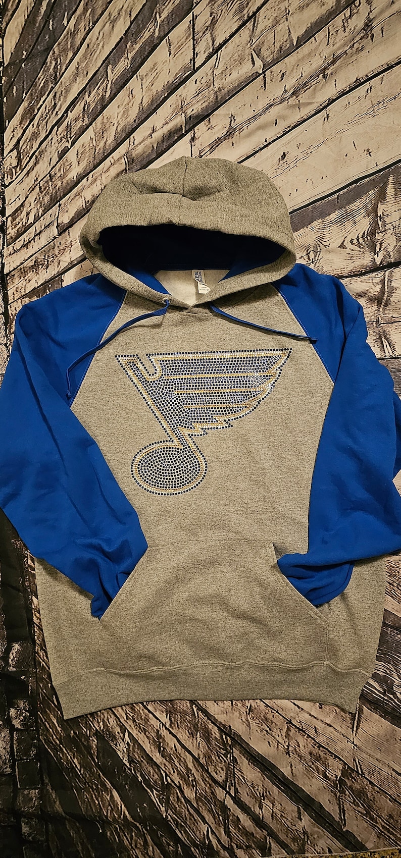 May include: Gray hoodie with blue sleeves and a hood. The front features a rhinestone design of the St. Louis Blues logo. The hoodie has a front pocket and a drawstring hood.