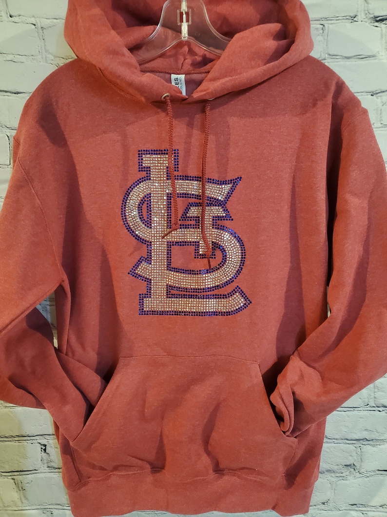 May include: Red hooded sweatshirt with a large rhinestone design of the St. Louis Cardinals logo in gold and purple.