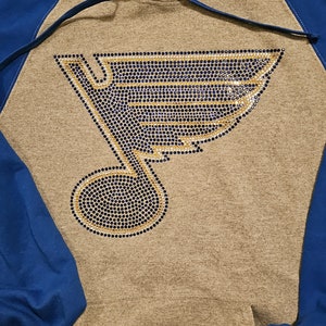 May include: Gray hoodie with blue sleeves and hood lining. The front features a rhinestone design of a musical note and a stylized wing in blue, gold, and silver. A drawstring is visible at the hood.