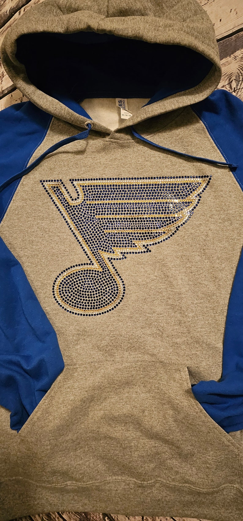 May include: Gray hoodie with blue sleeves and hood lining. The front features a rhinestone design of a musical note and a stylized wing in blue, gold, and silver. A drawstring is visible at the hood.