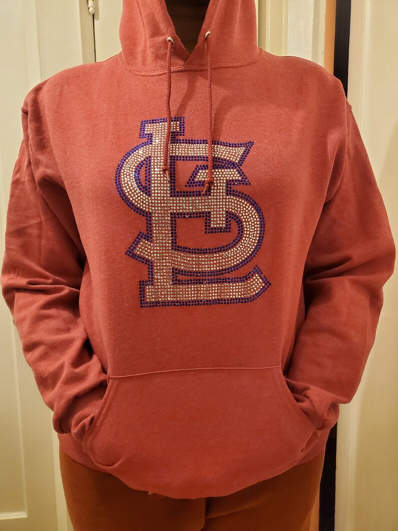 May include: A red hoodie with a large, sparkly, blue and gold "LS" logo on the front. The hoodie has a drawstring hood and a kangaroo pocket.