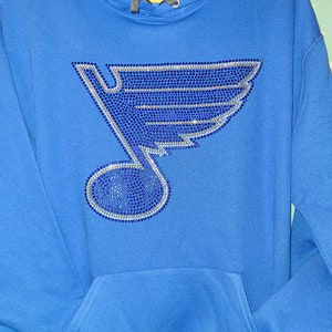 May include: A royal blue hoodie with a large rhinestone design of a hockey team's logo. The logo is a blue and silver note with a wing on top.