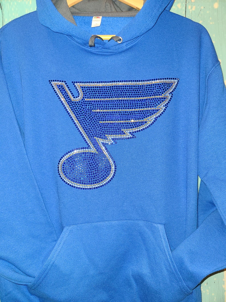 May include: A royal blue hoodie with a large rhinestone design of a hockey team's logo. The logo is a blue and silver note with a wing on top.