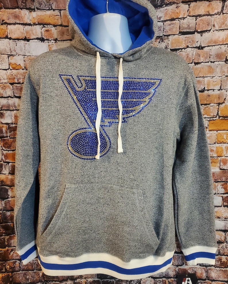 May include: Gray hoodie with a blue interior and a large blue and gold emblem on the front. The hoodie has a drawstring hood, a front pocket, and blue and white striped trim on the cuffs and hem.
