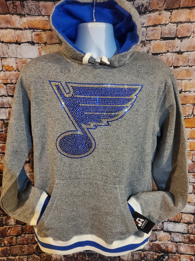 May include: Gray hoodie with a blue and gold rhinestone logo. The hoodie has a blue hood lining, white drawstrings, and a blue and white striped hem. The pockets and cuffs have white and blue accents. A tag is attached to the pocket.