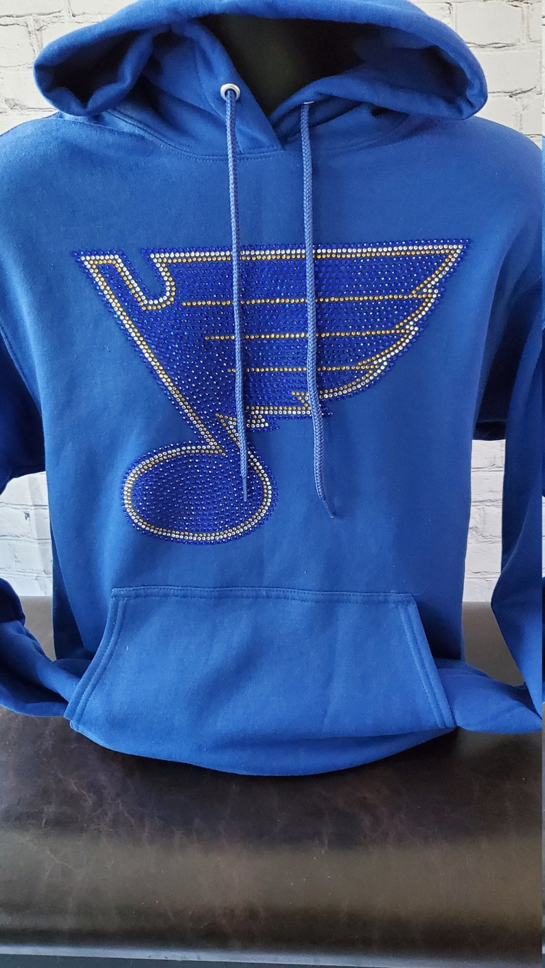 May include: A royal blue hoodie with a large, rhinestone-encrusted hockey team logo on the front. The logo is in the shape of a bird with wings and a hockey stick. The rhinestones are blue, gold, and silver.