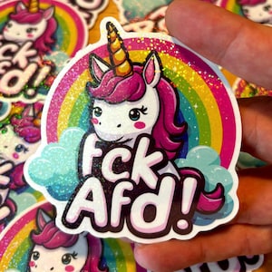 Könnte beinhalten: Ein weißes Einhorn mit einer pinken Mähne und einem Horn steht auf einem Regenbogen mit einer Wolke dahinter. Das Einhorn hält ein Schild mit der Aufschrift "FCK AFD!" Das Schild ist mit Glitzer bedeckt.