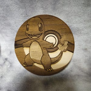 Könnte beinhalten: Ein runder Holzuntersetzer mit einem lasergravierten Design eines Charmander-Charakters aus dem Pokémon-Franchise. Der Untersetzer hat eine hellbraune Farbe mit detaillierten Schattierungen, und der Charakter lächelt. Das Design beinhaltet einen Pokéball.