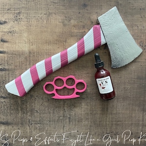 May include: A pink and white striped axe, a pink brass knuckle, and a small bottle of fake blood labeled "Fake Blood US Special Effects" on a wood background.