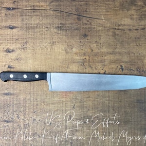 May include: A silver kitchen knife with a black handle. The knife is made of foam and is a prop for Halloween costumes. The text "WS Props & Effects" and "Foam Kitchen Knife Foam Michael Myers Knife" is written in white on a wooden surface.