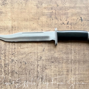 May include: A silver and black foam prop knife, resembling a hunting knife, on a wooden surface.