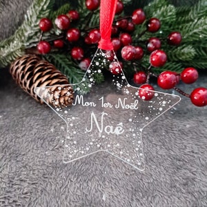 Peut inclure: Ornement de Noël transparent en forme d'étoile avec le texte "Mon 1er Noël Naé" et des détails de flocons de neige. L'ornement est suspendu à un ruban rouge. Le fond comprend des aiguilles de pin, des baies rouges et une pomme de pin.