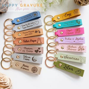 May include: A collection of fourteen rectangular leather keychains in various colors, each featuring a gold-tone metal ring and a gold-tone snap button. The keychains are engraved with diverse French phrases, names, and decorative icons.