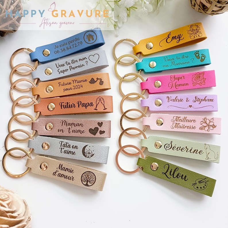 May include: A collection of fourteen rectangular leather keychains in various colors, each featuring a gold-tone metal ring and a gold-tone snap button. The keychains are engraved with diverse French phrases, names, and decorative icons.