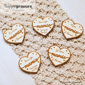 Peut inclure: Cinq plaques en bois en forme de cœur avec des surfaces claires et des bords brun foncé. Chaque plaque présente des textes gravés en français, notamment 'Bonne fête mamie', 'On t'aime mamie d'Amour', 'Super mamie d'Amour' et 'Je t'aime mamie d'Amour'.