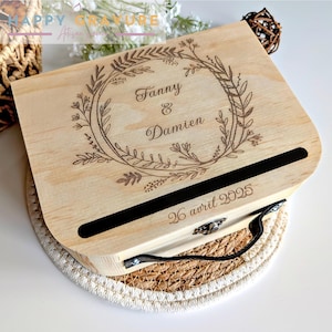Puede incluir: Una caja de madera de color claro con tapa abatible que presenta un dise&ntilde;o de corona floral grabado con l&aacute;ser. El centro de la corona muestra los nombres Fanny & Damien. Debajo de la corona, hay una ranura horizontal negra y la fecha 26 avril 2025.