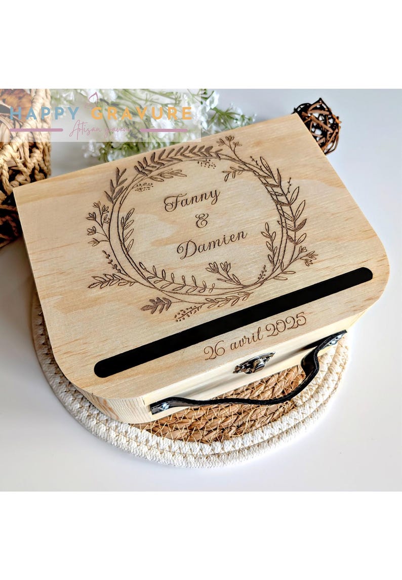 Puede incluir: Una caja de madera de color claro con tapa abatible que presenta un dise&ntilde;o de corona floral grabado con l&aacute;ser. El centro de la corona muestra los nombres Fanny & Damien. Debajo de la corona, hay una ranura horizontal negra y la fecha 26 avril 2025.