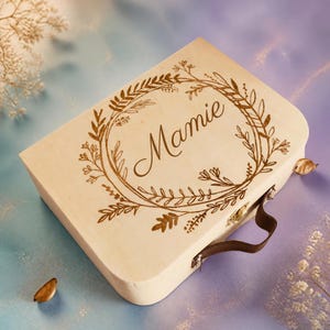 Puede incluir: Una maleta de madera de color claro con un asa marr&oacute;n oscuro y un cierre met&aacute;lico. La superficie superior presenta un dise&ntilde;o grabado con l&aacute;ser de una corona floral que rodea la palabra Mamie en letra cursiva, centrada en la tapa.