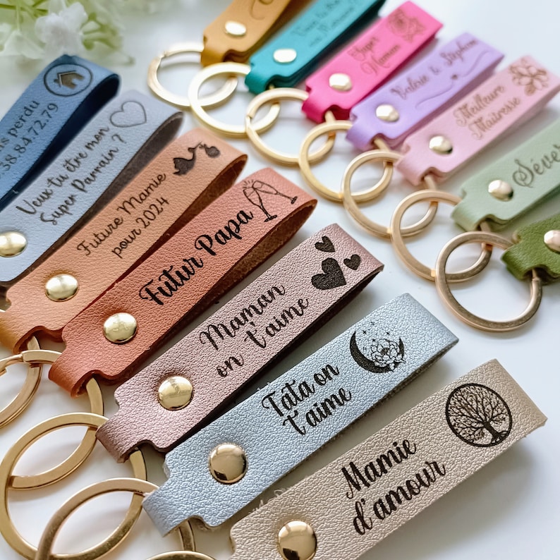 May include: A collection of multi-colored faux leather keychains featuring gold-tone metal rings and rivets. Each strap is engraved with various French phrases, names, and icons such as hearts, a tree, a moon, and champagne glasses.