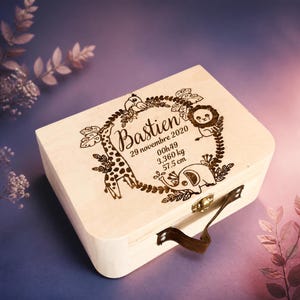 Puede incluir: Una caja de madera clara rectangular con un cierre met&aacute;lico y un asa marr&oacute;n. La tapa presenta un dise&ntilde;o grabado con l&aacute;ser de una corona con una jirafa, un le&oacute;n, un elefante y un p&aacute;jaro, adem&aacute;s del texto Bastien, 29 novembre 2020, 00h49, 3,360 kg, 57,5 cm.