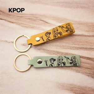 May include: Two keychains with metal rings and leather-like straps in mustard yellow and sage green. Each strap features a silver-tone snap button and a black line-art illustration of three stylized female figures. The text KPOP is visible.