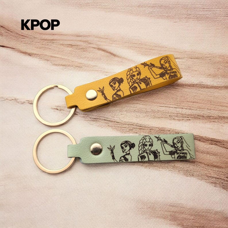 May include: Two keychains with metal rings and leather-like straps in mustard yellow and sage green. Each strap features a silver-tone snap button and a black line-art illustration of three stylized female figures. The text KPOP is visible.