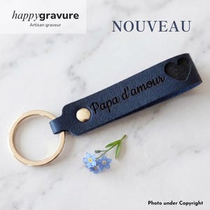 May include: A dark blue leather keychain featuring a gold-toned metal ring and a gold rivet. The strap is engraved with the text Papa d'amour and includes a heart-shaped cutout. A small sprig of light blue forget-me-not flowers lies nearby.