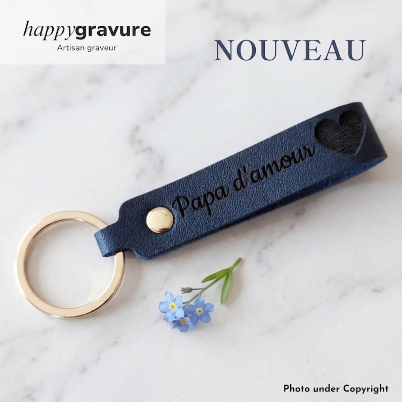 May include: A dark blue leather keychain featuring a gold-toned metal ring and a gold rivet. The strap is engraved with the text Papa d'amour and includes a heart-shaped cutout. A small sprig of light blue forget-me-not flowers lies nearby.
